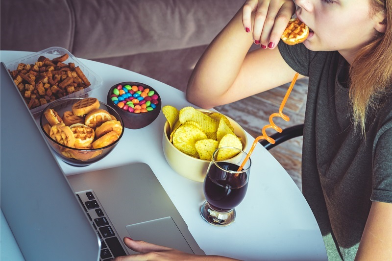 Girl works at a computer and stress eats. Unhealthy food: chips, crackers, candy, waffles, soda.