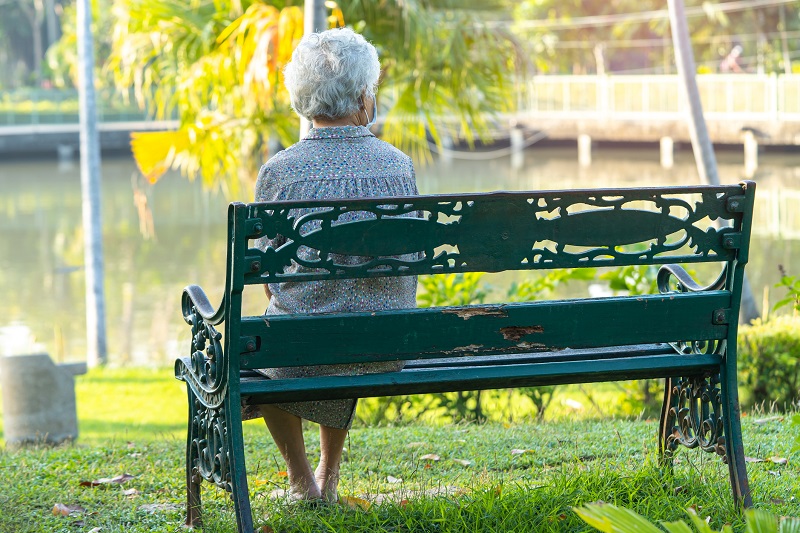 Older woman sitting on a park bench near the water as the sun goes down.